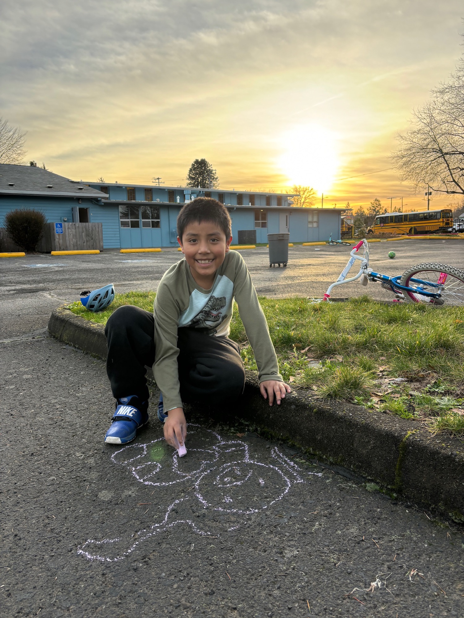 A boy smiling while drawing with chalk on the pavement at sunset